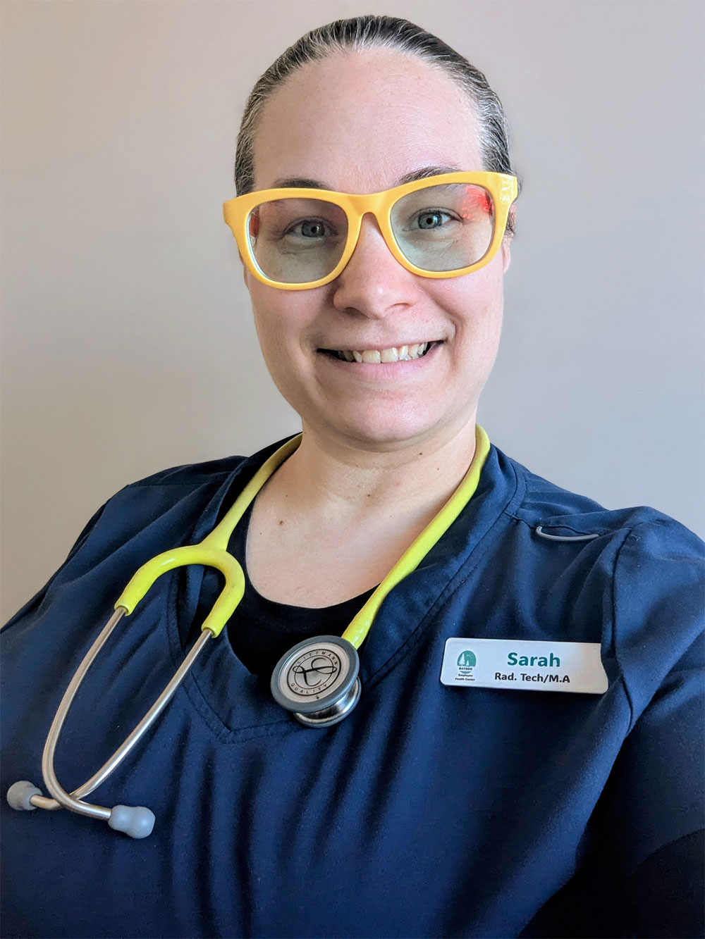 An x-ray technician wears her yellow fun glasses, and blue scrubs smiling against a gray wall.